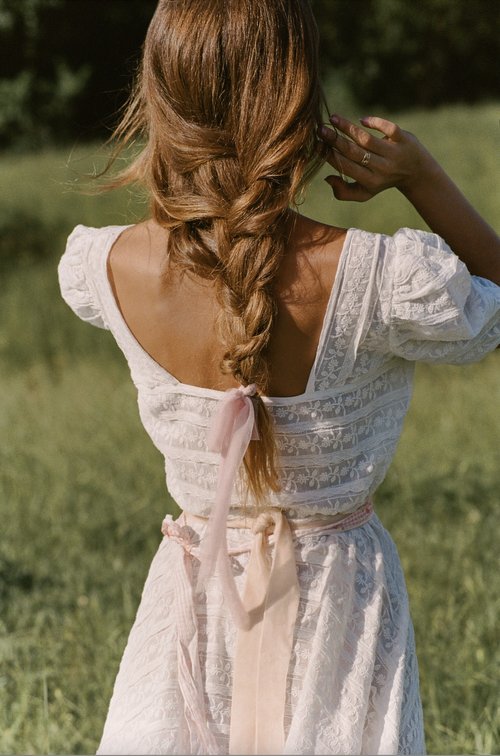 Woman in white lace dress with a braided hairstyle tied with a pink ribbon outdoors in a grassy meadow.