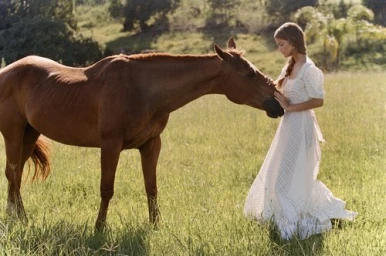 A woman in a white dress standing in a grassy field, gently touching the head of a brown horse.