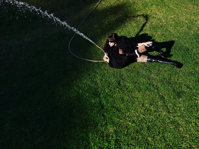 Woman in black dress and boots lies on grass, being sprayed with water from a garden hose in an overhead, outdoor shot.