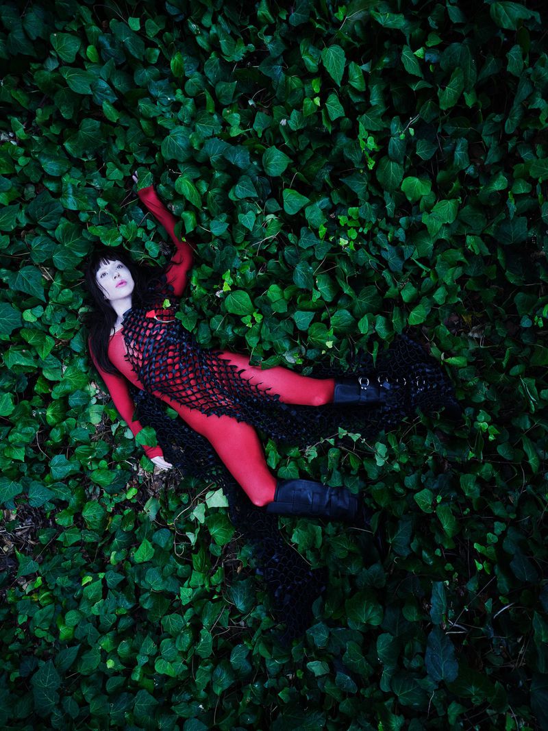 Adult female model in full red bodysuit with black mesh overlay posing in lush green ivy for a fashion or editorial portrait.