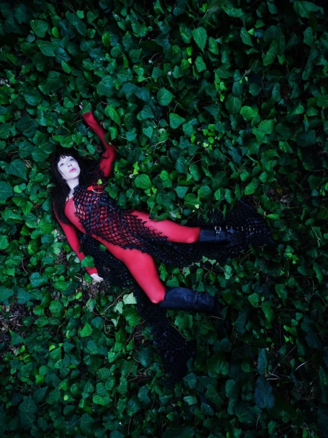 Adult female model in full red bodysuit with black mesh overlay posing in lush green ivy for a fashion or editorial portrait.