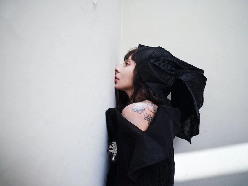 Young woman in stylish black outfit and dramatic headpiece poses in corner of room, looking away, tattoo visible on her shoulder.
