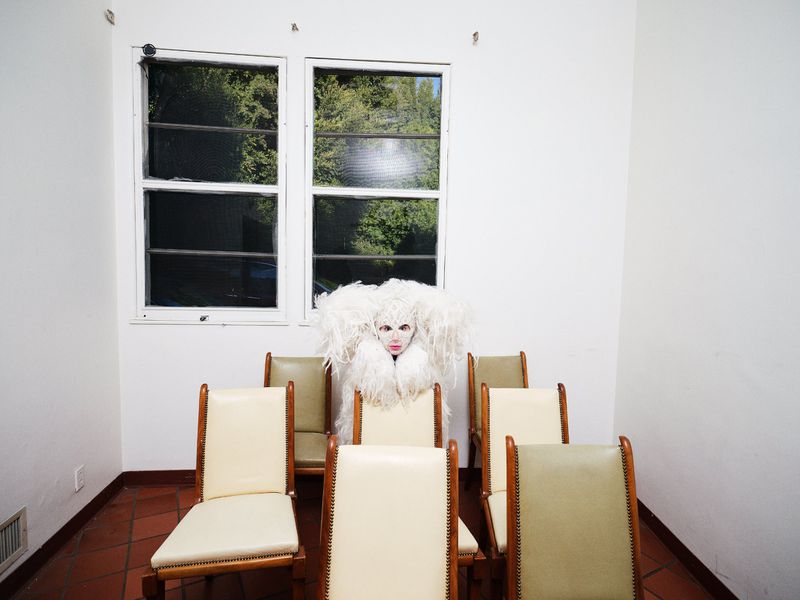Wide shot of a performer in a white feathered costume and mask sitting behind cream-colored chairs in an empty white room with red tile.