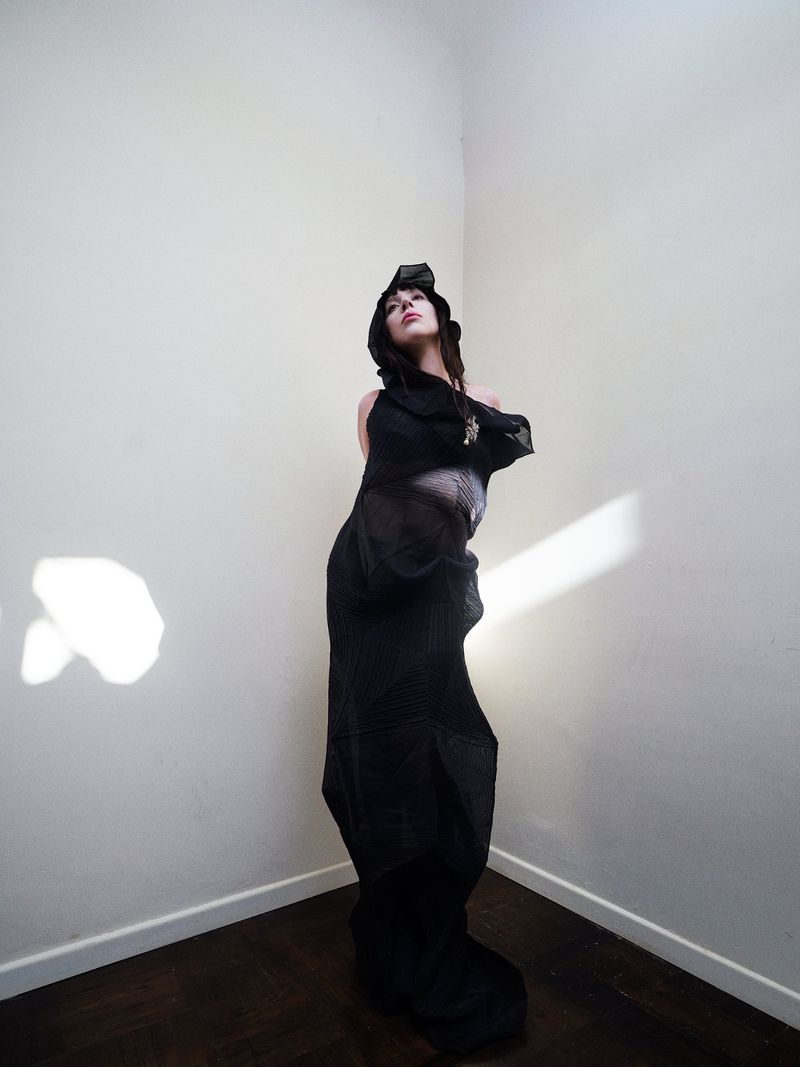 Woman in dramatic hooded outfit stands in a corner with dark wood floors. Bright, diffused lighting highlights the scene.