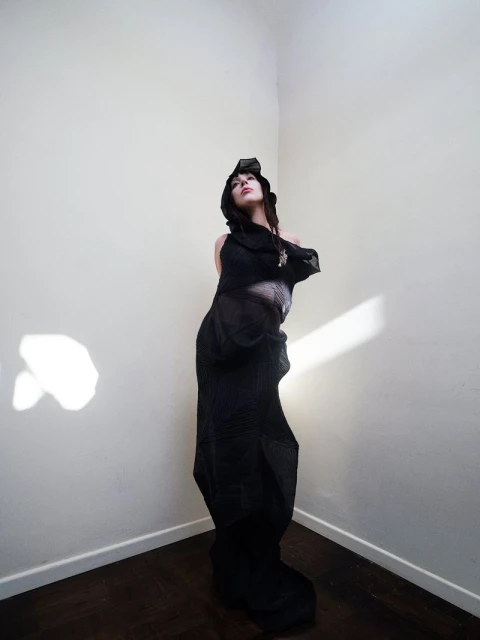 Woman in dramatic hooded outfit stands in a corner with dark wood floors. Bright, diffused lighting highlights the scene.