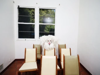 Lady Gaga in an elaborate white feather costume seated amidst chairs in a room for Elle U.S. February 2025.