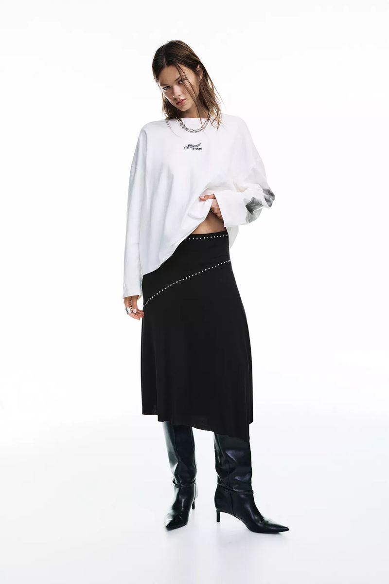 A woman poses in a white long-sleeved shirt, black skirt, and black knee-high boots against a bright white background in a studio shot.