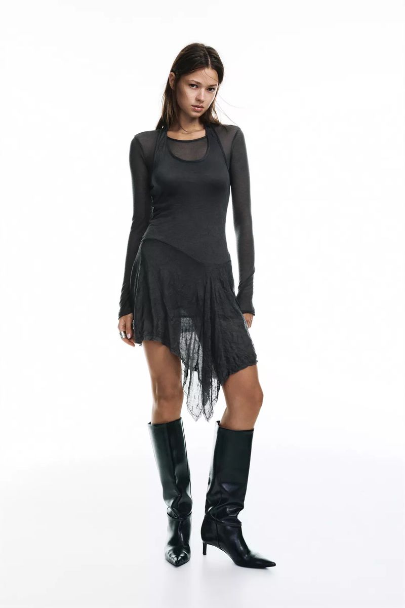 Full-length studio shot of a young woman posing in a dark gray, long-sleeved top and asymmetric skirt, paired with black boots against a white backdrop.