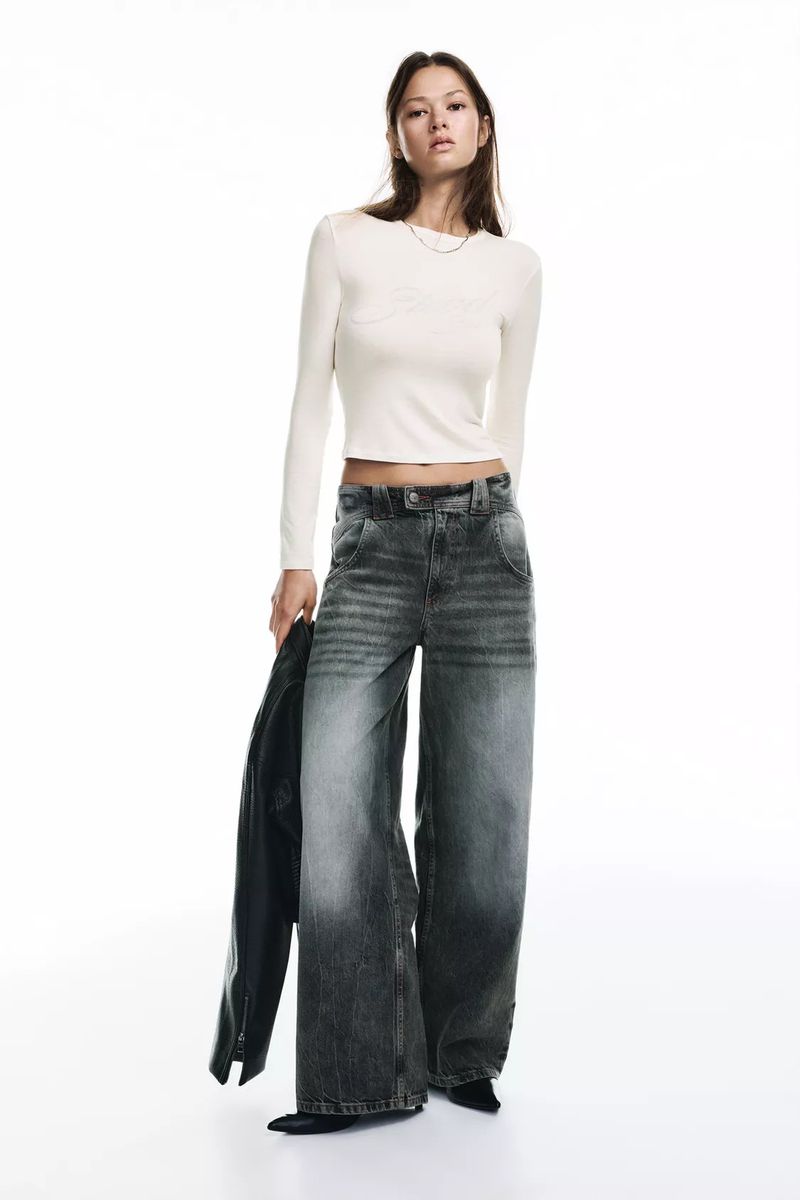 Fashionable woman in a white top and wide-leg jeans holding a leather jacket against a bright backdrop, studio shot.