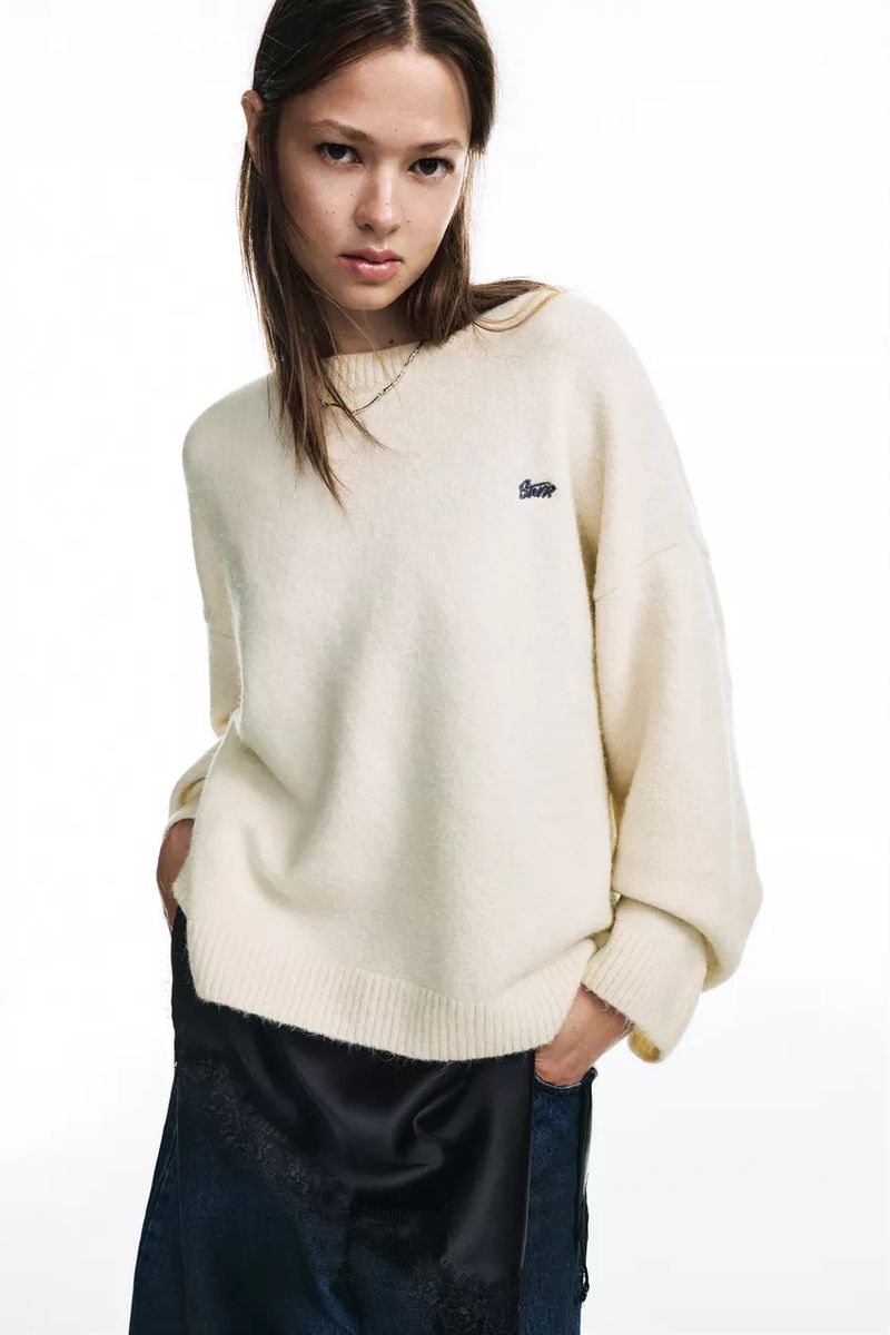 Woman in oversized cream sweater and dark skirt poses in a studio setting against a white backdrop, hands in her pockets.