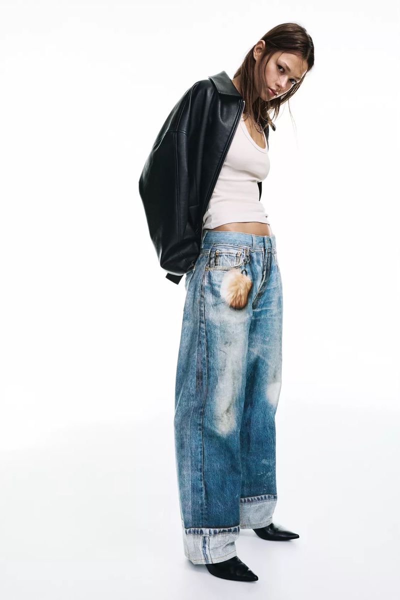 Fashionable young adult woman in baggy jeans and a leather jacket posing in a studio with a clean white backdrop and studio lighting.