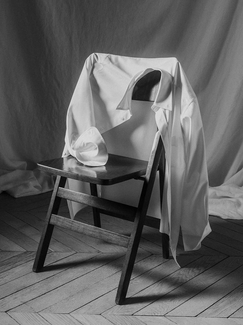 Monochrome still life featuring a white shirt and hat draped on a dark wooden chair against a neutral fabric backdrop indoors.