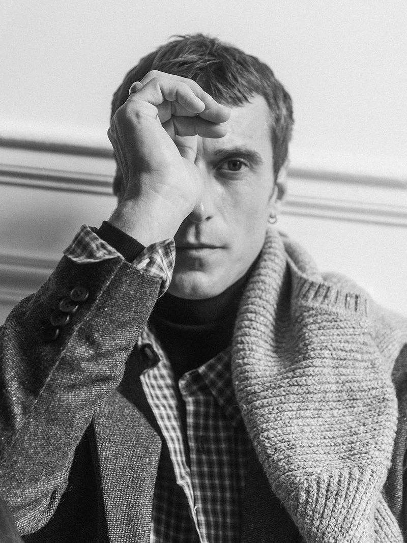Clement Chabernaud posing with a hand over his forehead wearing a layered tweed blazer, knit sweater, and plaid shirt in black and white.