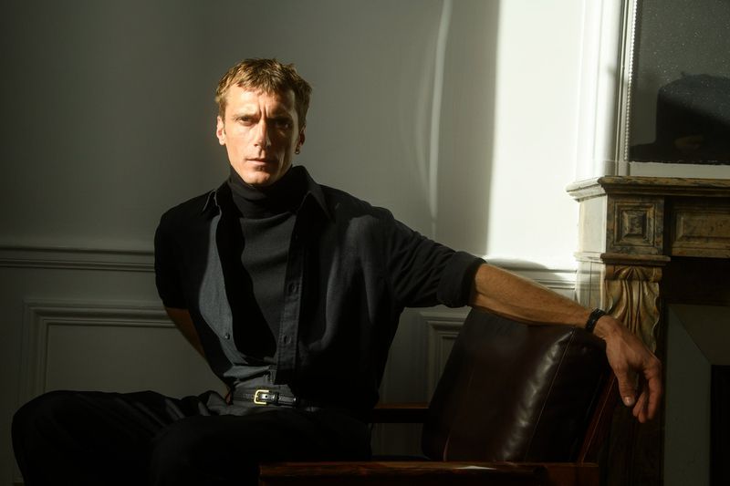 Portrait of an adult male sitting in a chair, wearing a black turtleneck and open shirt in an indoor setting with natural light.