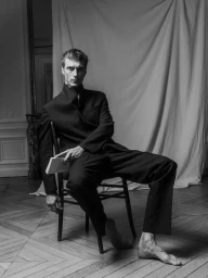 Model Clement Chabernaud in a dark suit holding a book while seated on a chair against a draped backdrop for Zara Man