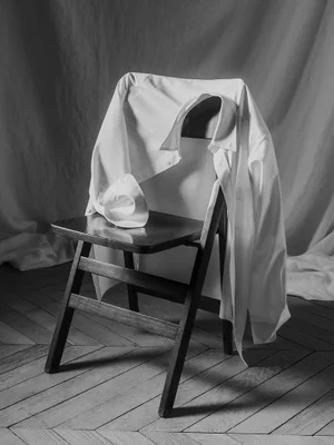 A white collared dress shirt draped over the back of a dark wooden folding chair on a herringbone parquet floor in black and white.