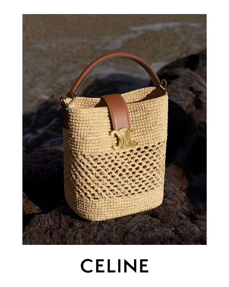 A light brown woven bucket bag with a brown leather handle and strap detail sits on dark rocks with a blurred ocean background.