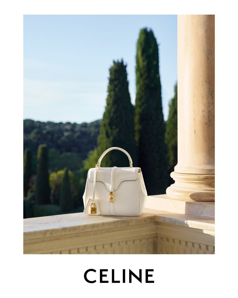 Luxury white leather Celine handbag with gold hardware displayed outdoors on a stone ledge with trees and blue sky background.