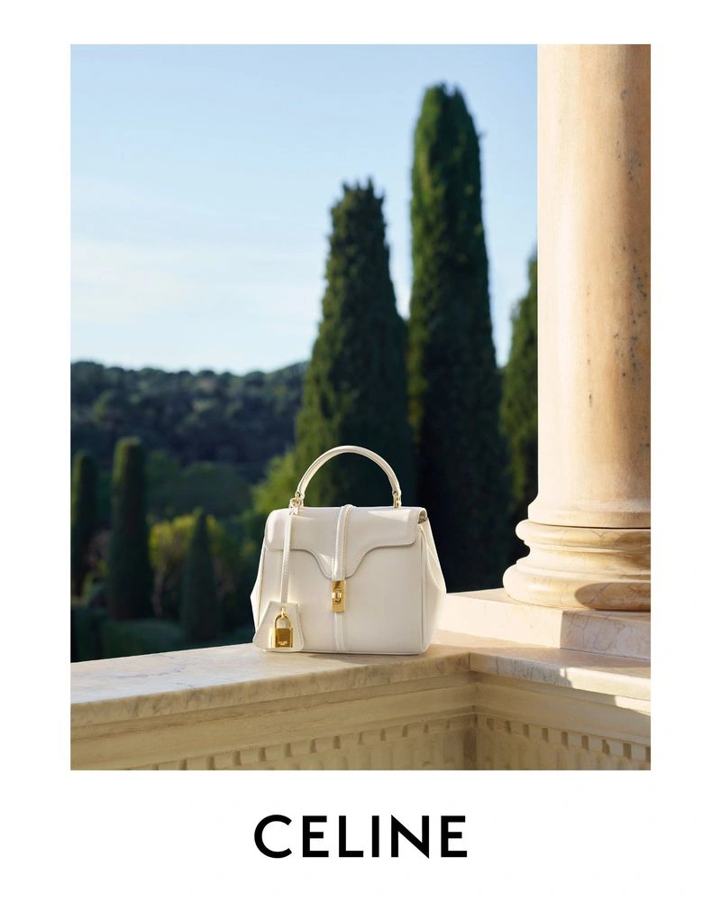 White leather Celine handbag with gold hardware resting on a sunlit stone balustrade beside a column with cypress trees in the background.