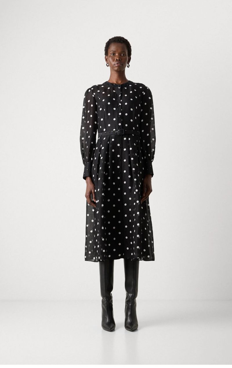 A Black woman model stands in a photo studio, wearing a long-sleeved black midi dress with white polka dots and black leather boots.