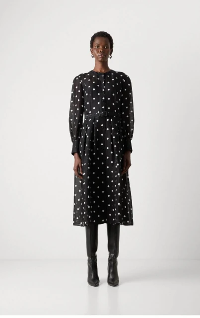 A Black woman model stands in a photo studio, wearing a long-sleeved black midi dress with white polka dots and black leather boots.