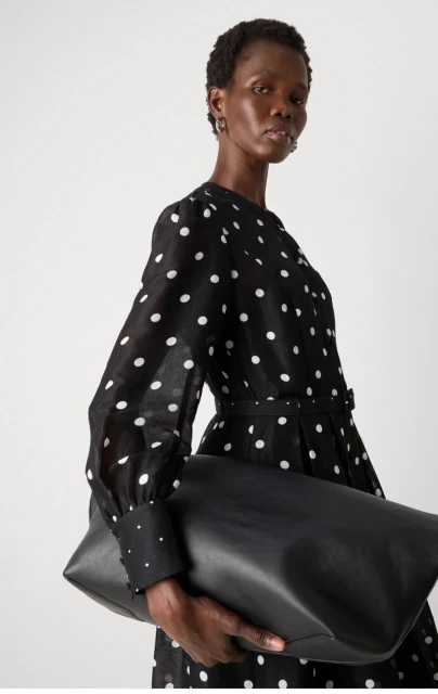 Woman wearing a black and white polka dot dress holds a black leather clutch bag against a plain white background.