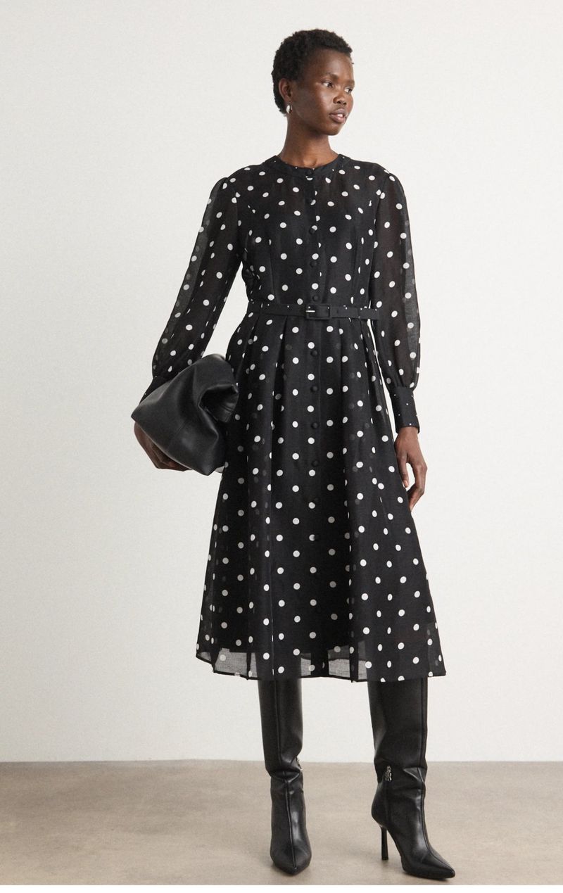 An African American woman poses in a black and white polka dot dress, accessorized with black knee-high boots and a black leather bag.