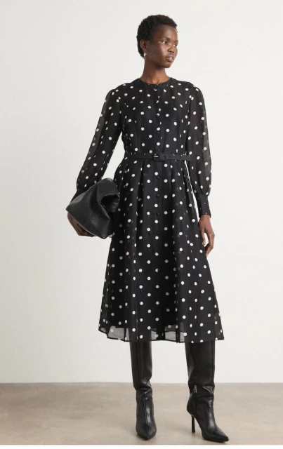 An African American woman poses in a black and white polka dot dress, accessorized with black knee-high boots and a black leather bag.