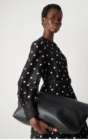 Model Hakima Athuai in a black polka dot dress holding an oversized black leather clutch bag against a light studio backdrop.