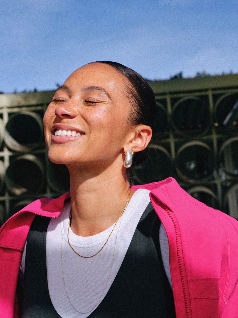 Smiling adult female with freckles, wearing a pink jacket, white t-shirt, and silver hoop earrings, standing outside on a sunny day.