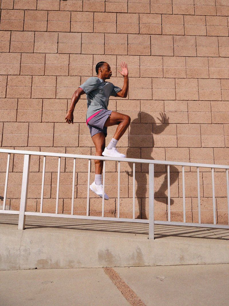 Athletic man steps over a metal railing outside; wearing athletic shorts and a t-shirt. The background features a brick wall.