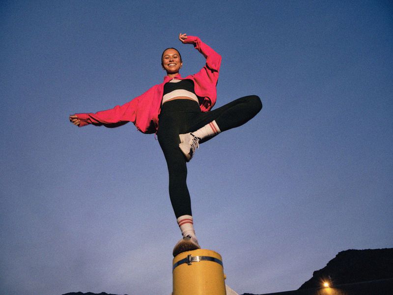 Young woman in athletic wear balances on a yellow post against a twilight sky, striking a dynamic pose with arms outstretched.