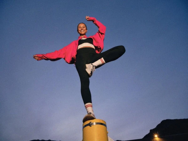 Young woman in athletic wear balances on a yellow post against a twilight sky, striking a dynamic pose with arms outstretched.