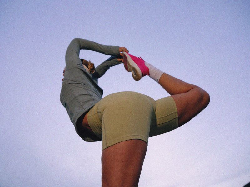 Female athlete in activewear performing a standing yoga pose against a light blue sky, showcasing fitness and flexibility.