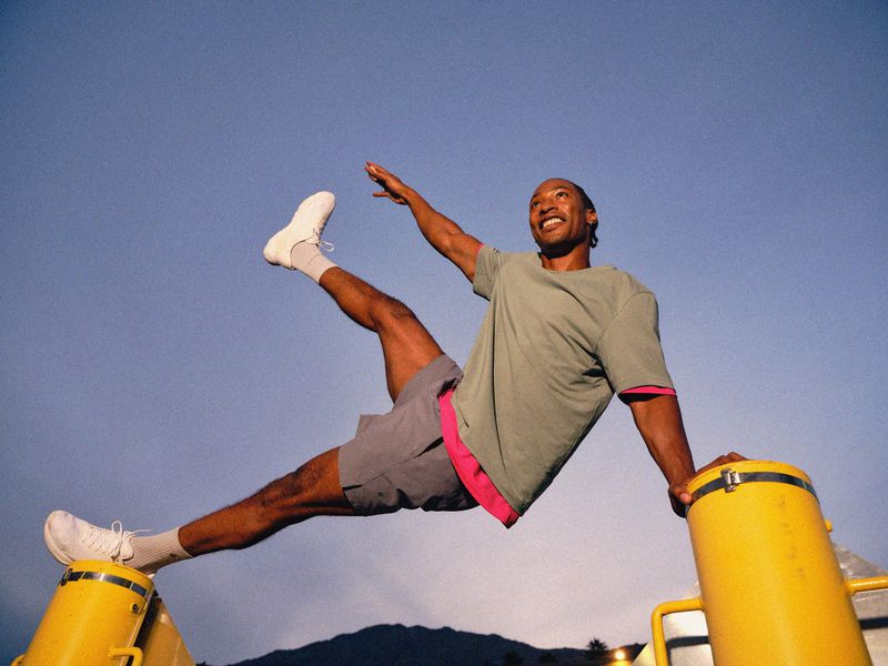 Athletic African American man in sportswear balances on yellow equipment against a blue sky, showcasing fitness and flexibility.