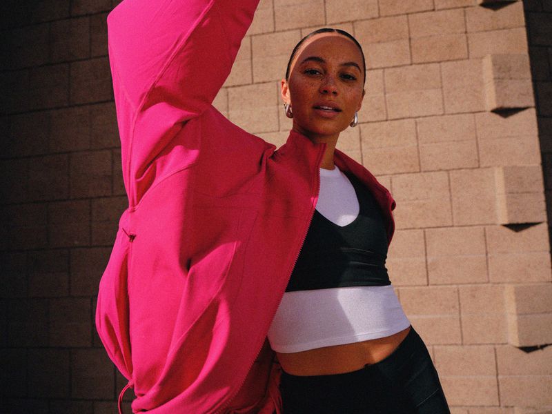 Woman posing in a bright pink athletic jacket, black sports bra, and white crop top against a cinder block wall.