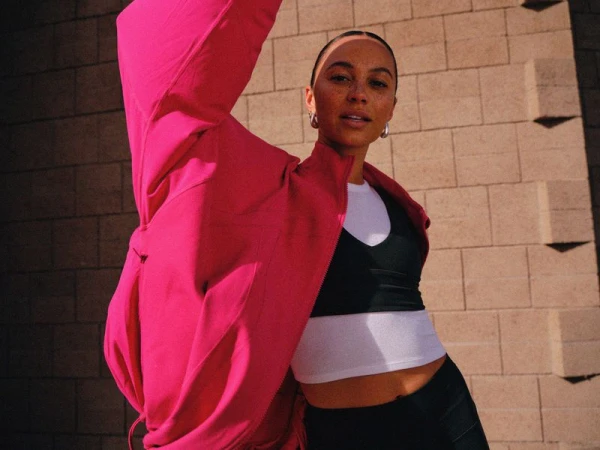 Woman posing in a bright pink athletic jacket, black sports bra, and white crop top against a cinder block wall.