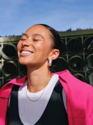 Close-up portrait of a smiling woman wearing a vibrant pink zip jacket and silver teardrop earring in direct sunlight.
