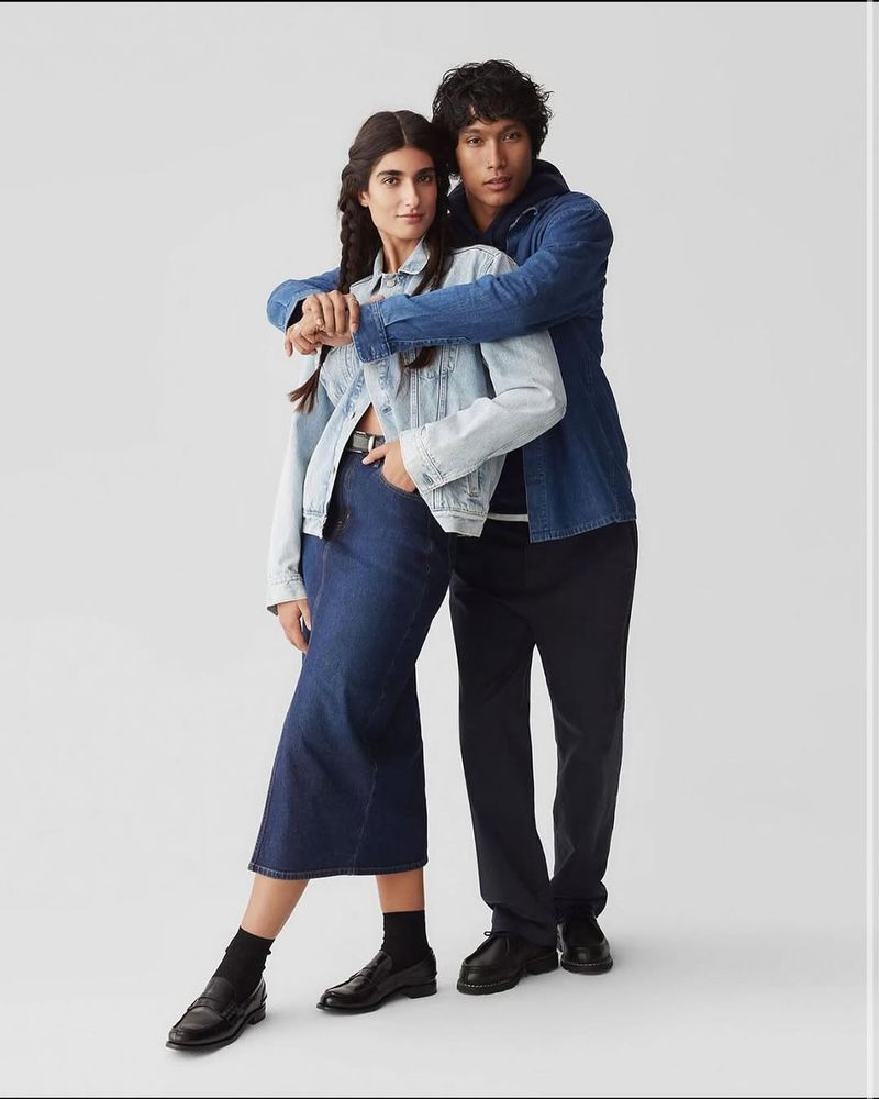 A stylish interracial couple poses in coordinating denim outfits against a neutral backdrop for a fashion editorial shoot.