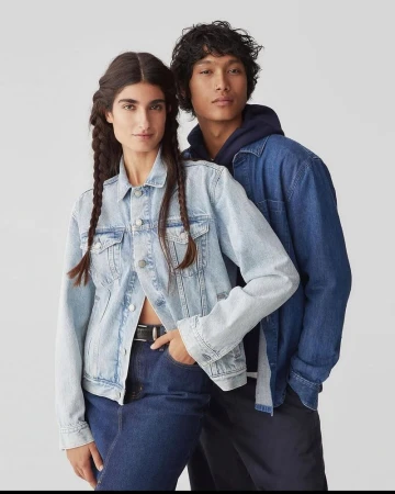 A woman in a light-wash denim jacket and a man in a blue denim shirt over a navy hoodie posing against a neutral studio backdrop.