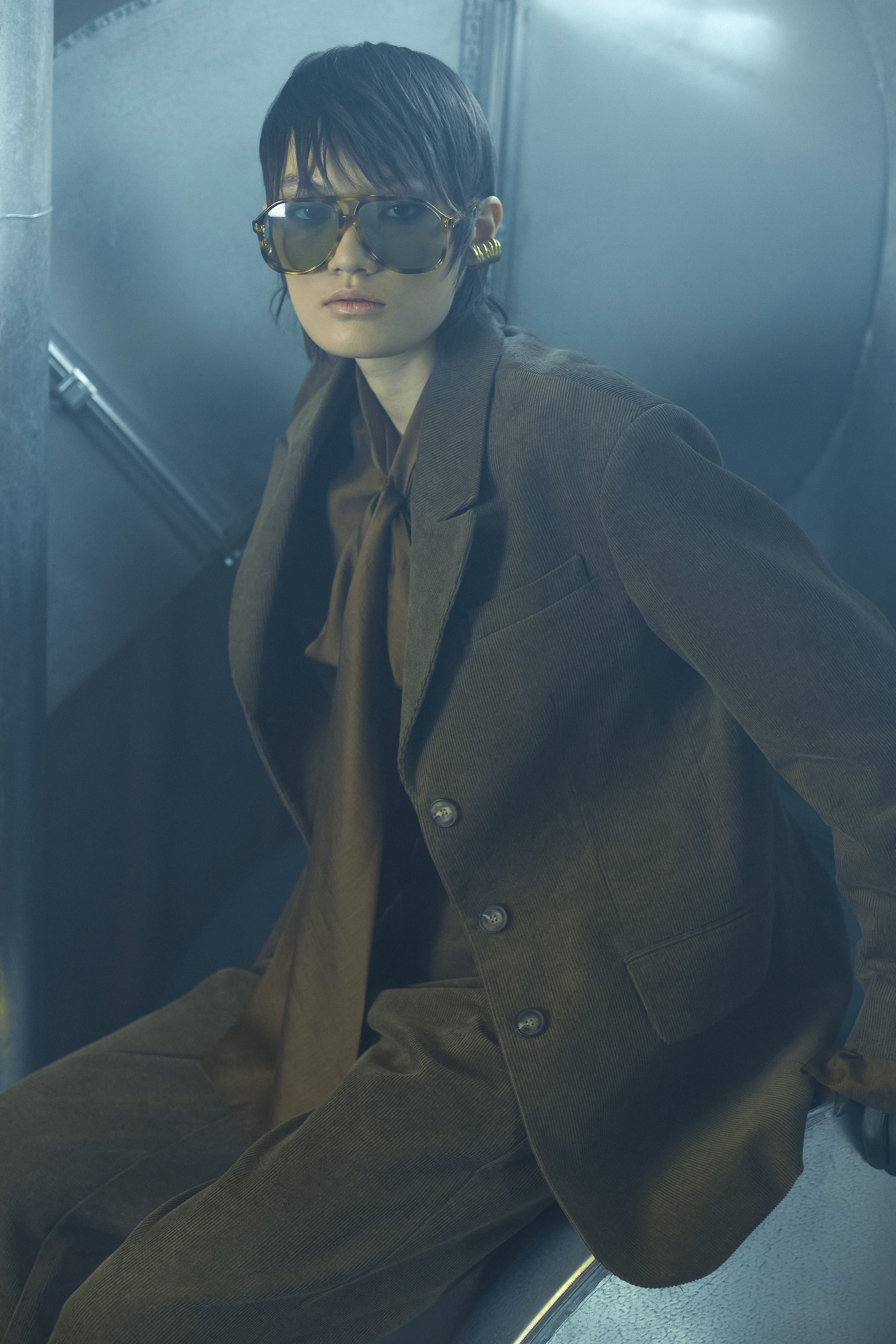 Woman in olive green suit and tortoise shell sunglasses sits in front of a textured metal backdrop in a studio setting.