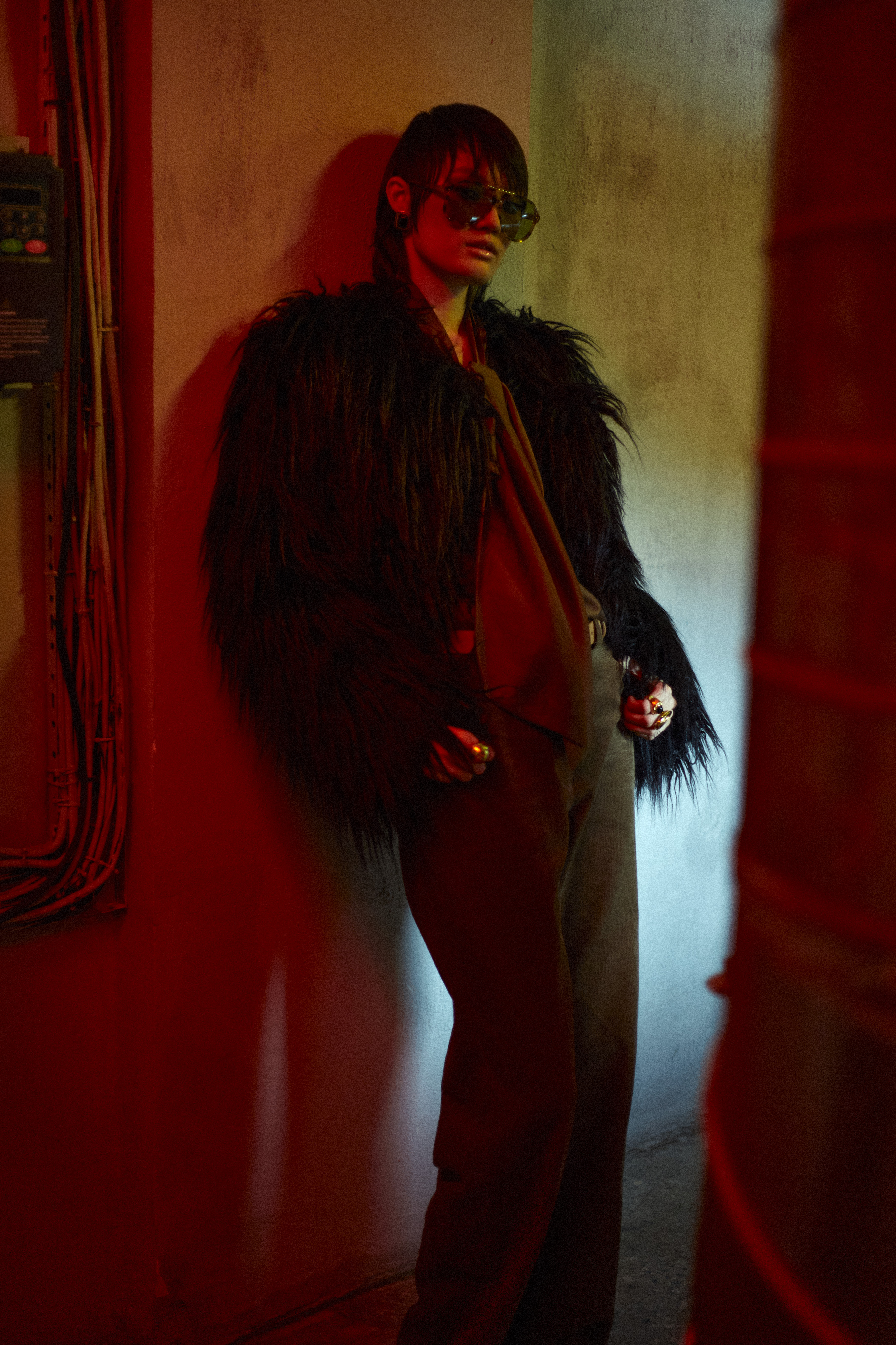 Stylish adult posing in a fur jacket, sunglasses, and pants against an indoor wall with dramatic red and blue lighting.