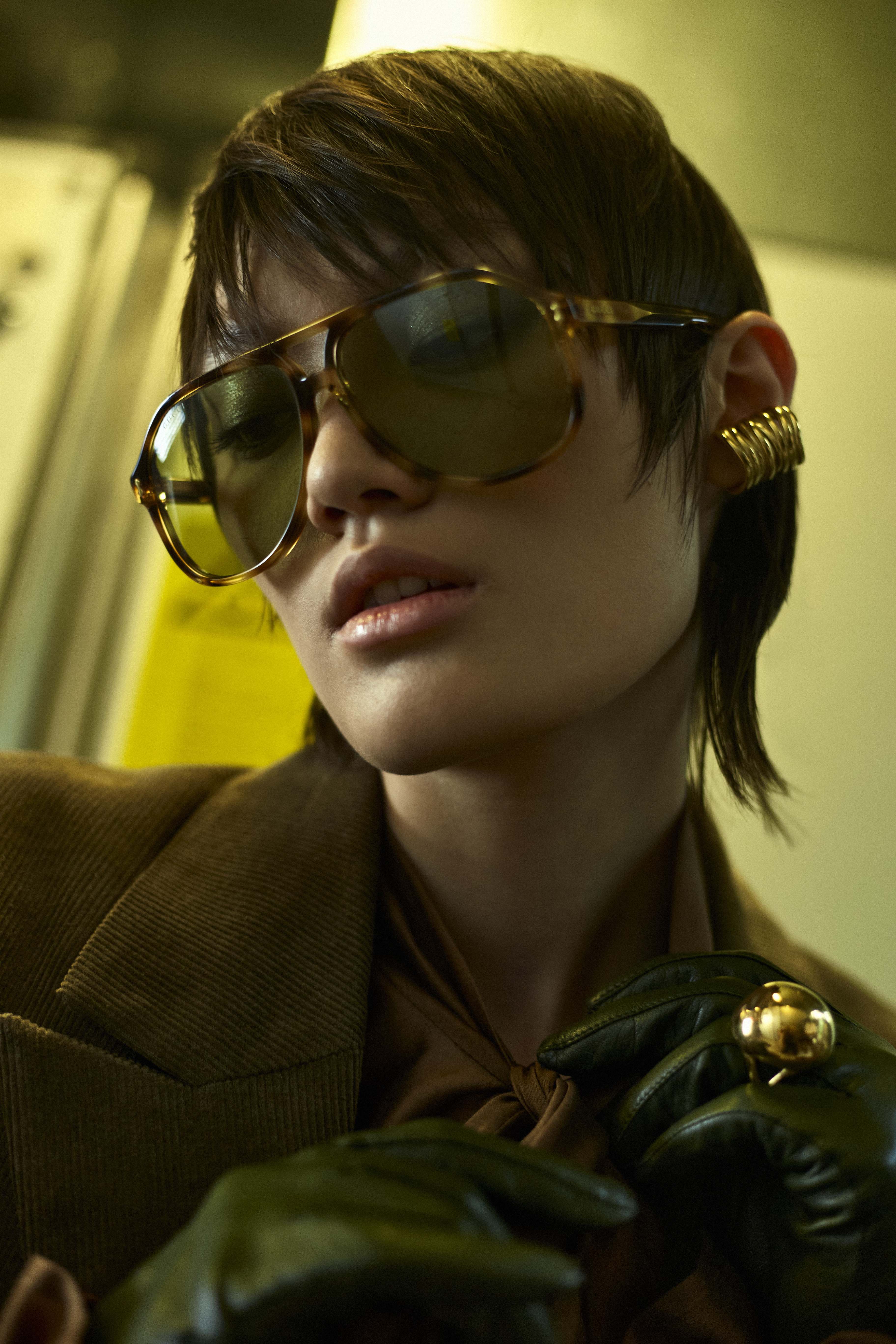 Close-up fashion portrait of a young adult woman with an olive green jacket, sunglasses, and ear cuff jewelry in a subway car.
