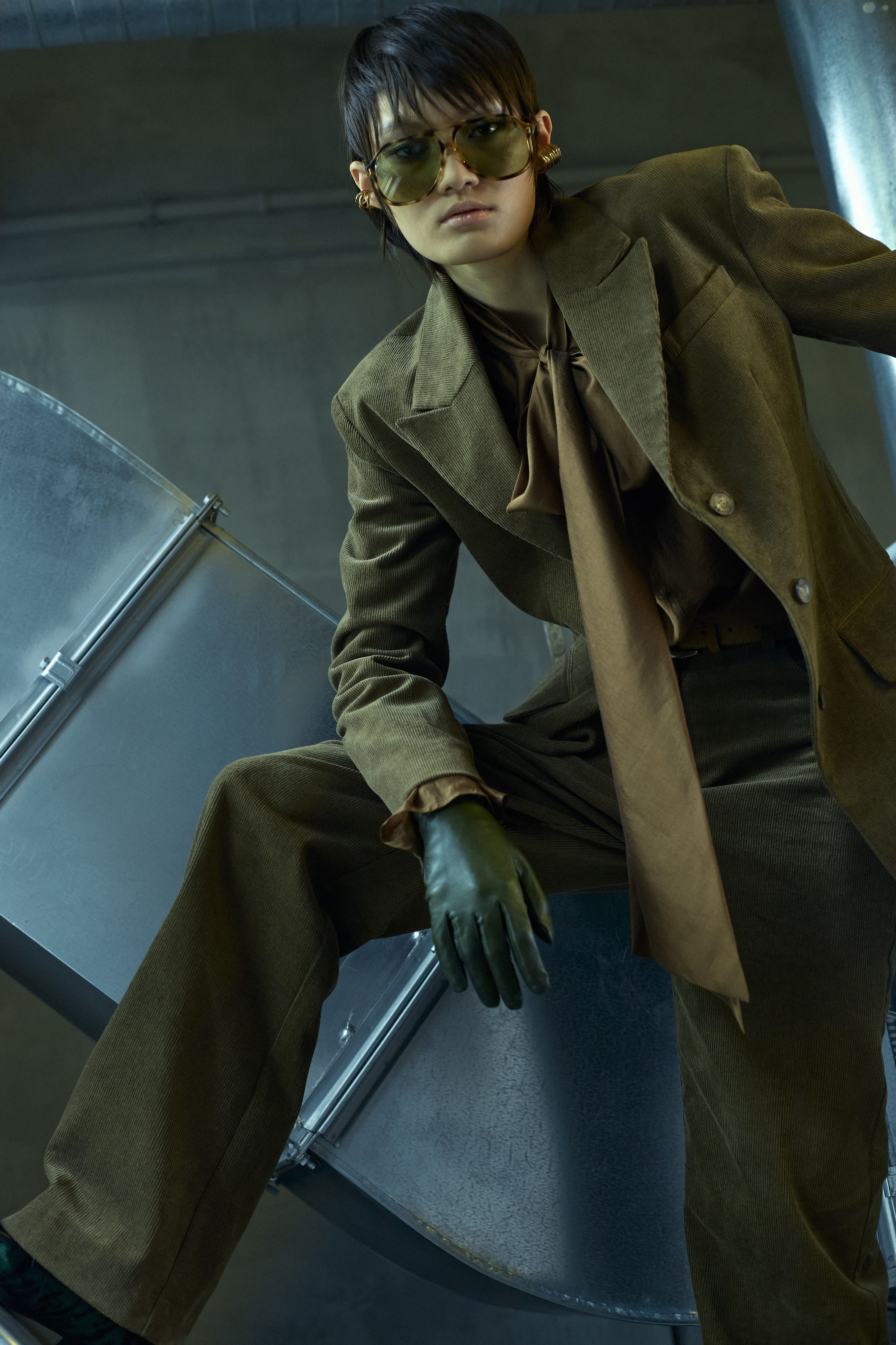 Stylish woman in a corduroy suit and tinted glasses sits on a metal ventilation system in an industrial interior, fashion editorial shot.