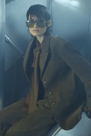 Model in brown blazer and tinted sunglasses seated inside a metallic industrial setting with cool hazy blue lighting.