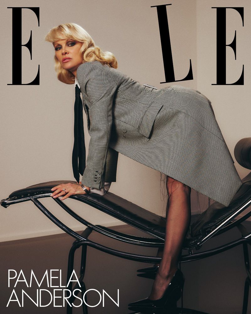 Pamela Anderson poses in a grey blazer suit, black tie and heels in front of the ELLE logo on a magazine cover; she leans on a black chair.