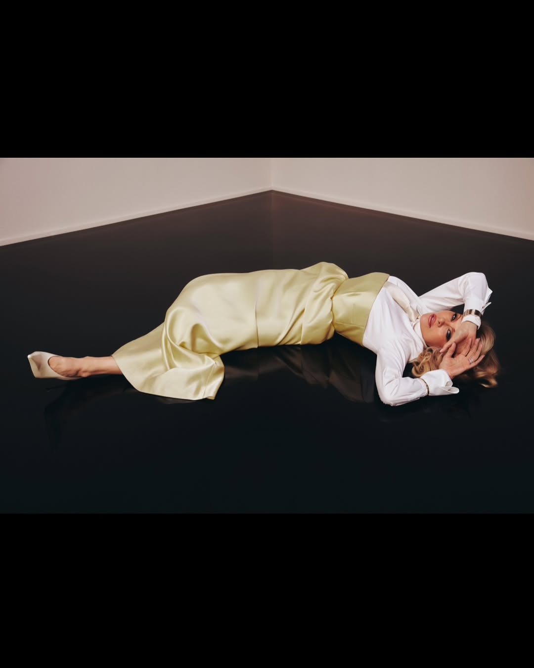 Adult woman in a light yellow satin outfit and white shirt lying on a black floor in a corner with neutral walls.