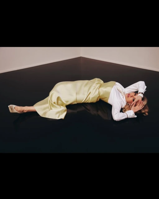 Adult woman in a light yellow satin outfit and white shirt lying on a black floor in a corner with neutral walls.