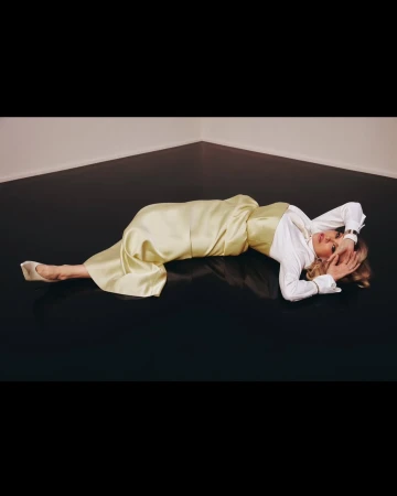 Adult woman in a light yellow satin outfit and white shirt lying on a black floor in a corner with neutral walls.