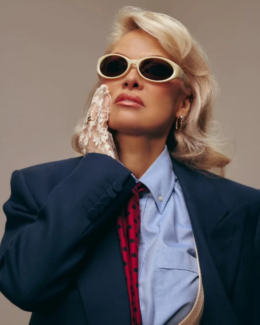 Stylish woman with blonde wavy hair and sunglasses poses in a blue blazer and light-blue shirt against a neutral background.